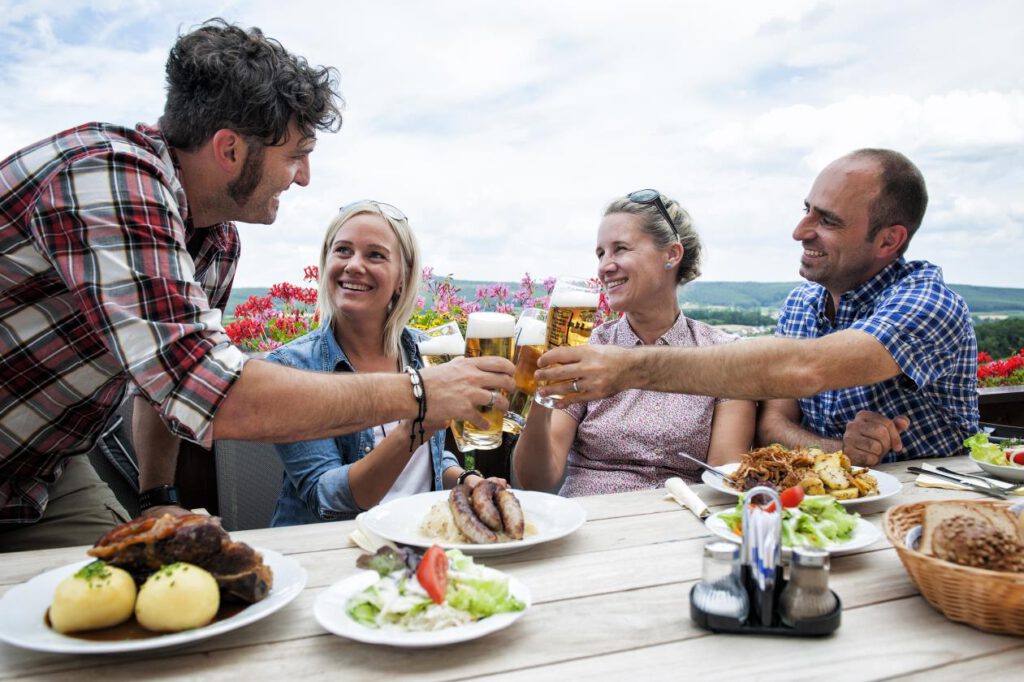 Auf dem Bild sind 4 Personen beim Essen zu sehen. es stehen verschiedene fränkische Gerichte wie Schäuferle und Bratwurt mit Sauerkraut auf dem Tisch. Die 4 Personen stoßen jeweils mit einem Glas Bier an